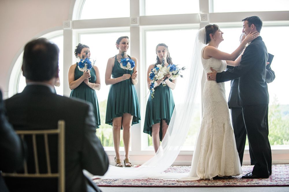 Bride walks down a path with two men, holding a yellow bouquet. Fountain and building in the background.