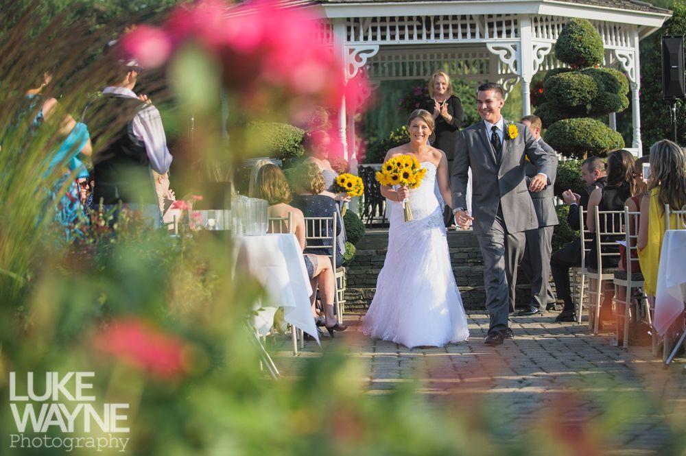 Newlyweds walk down aisle holding hands, sunflowers, gazebo in background.