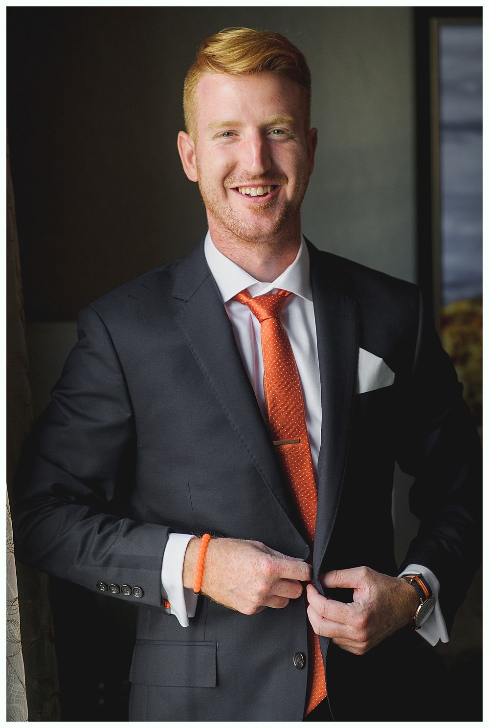 Man in a dark suit with an orange tie, smiling, adjusting his jacket.