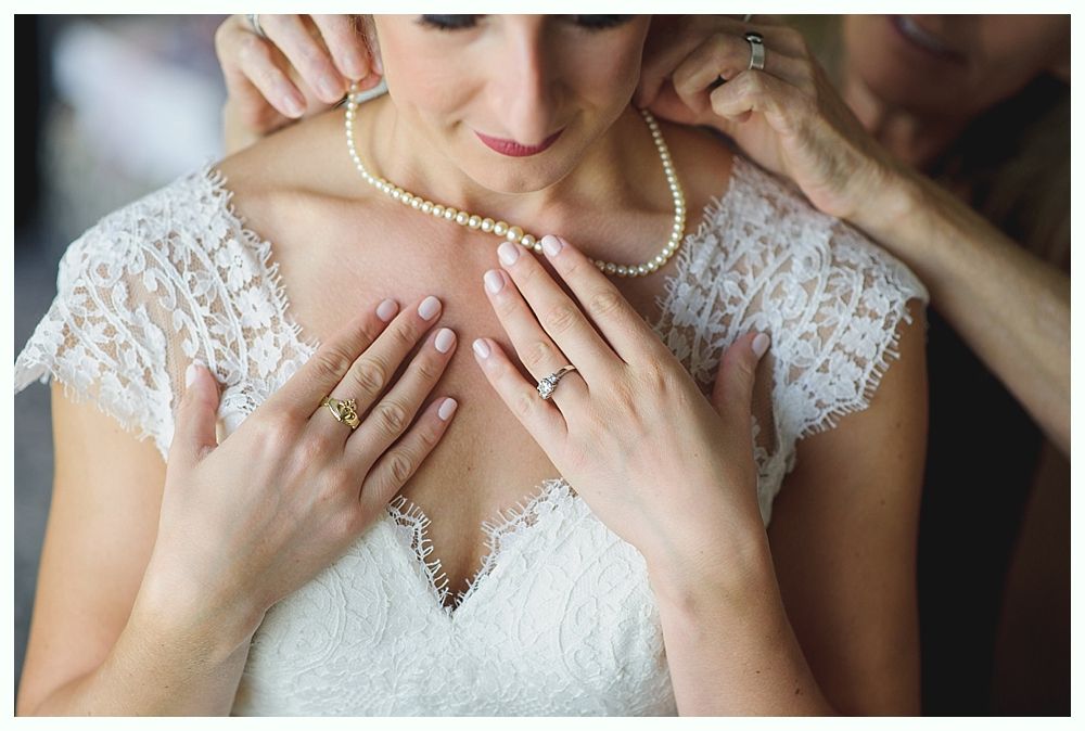 Bride in a white lace dress having pearl necklace clasped by another person's hands.