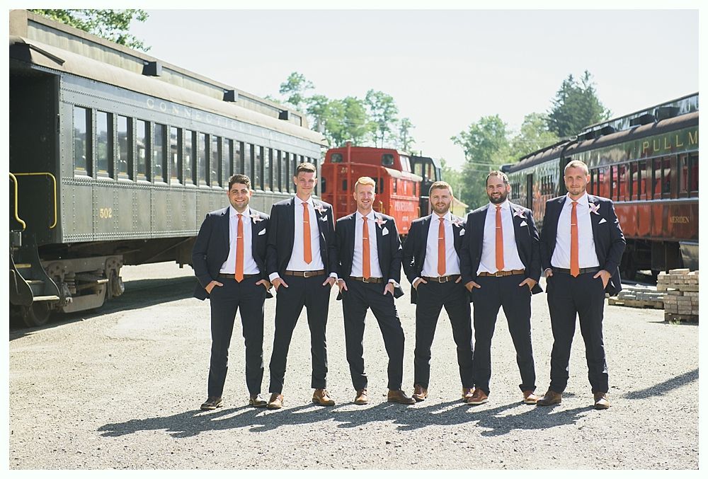 Six men in suits and orange ties pose near train cars, hands in pockets.