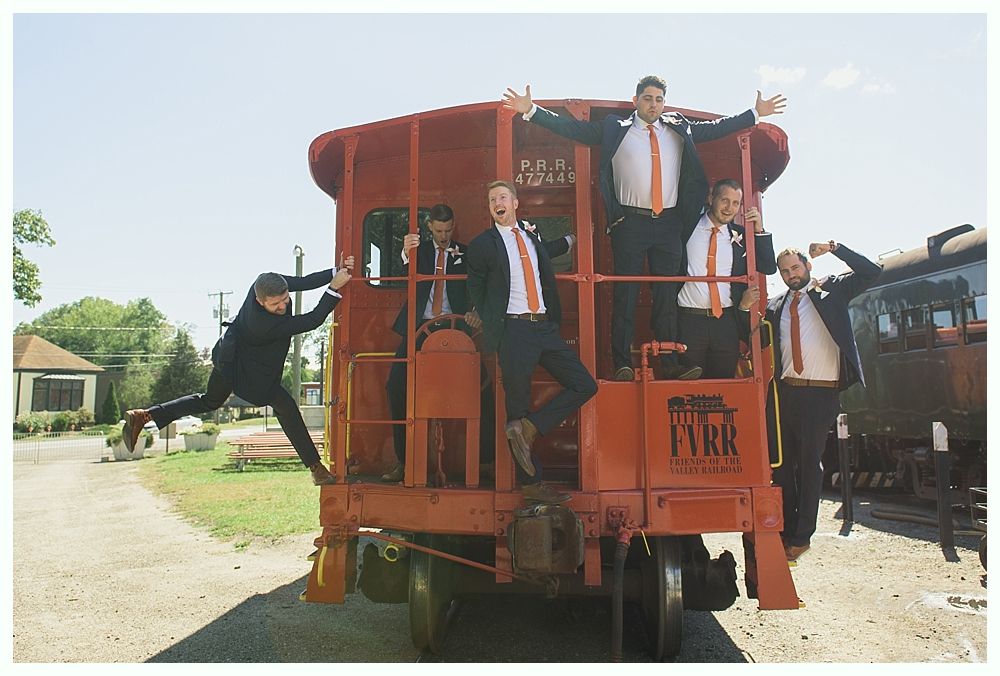 Men in suits posing on a red train caboose; arms raised, excited expressions. Outdoors on a sunny day.