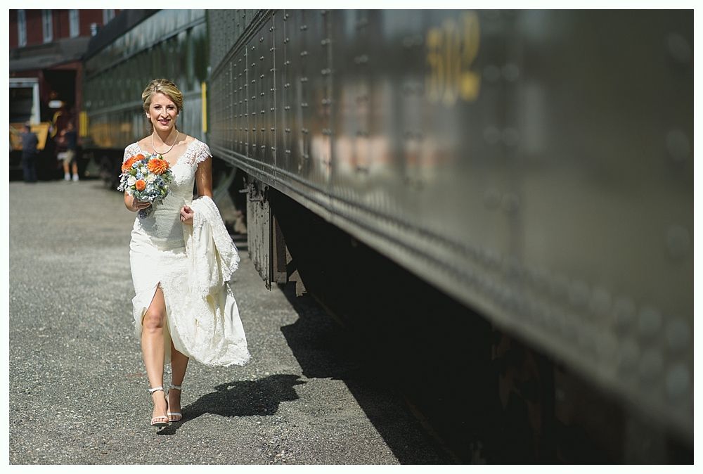 Bride in wedding dress, holding bouquet, walks past train car; smiling, outdoor setting.