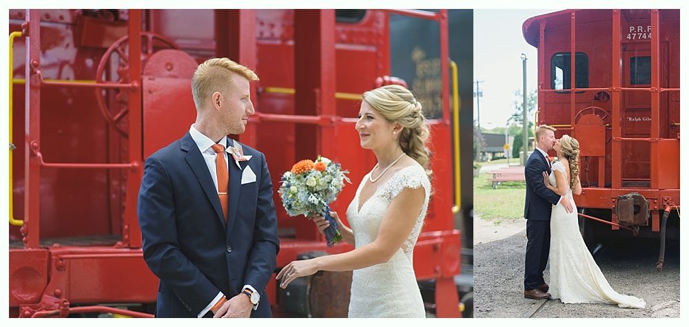 A newly married couple poses by a red train. The bride holds flowers and the groom kisses her.