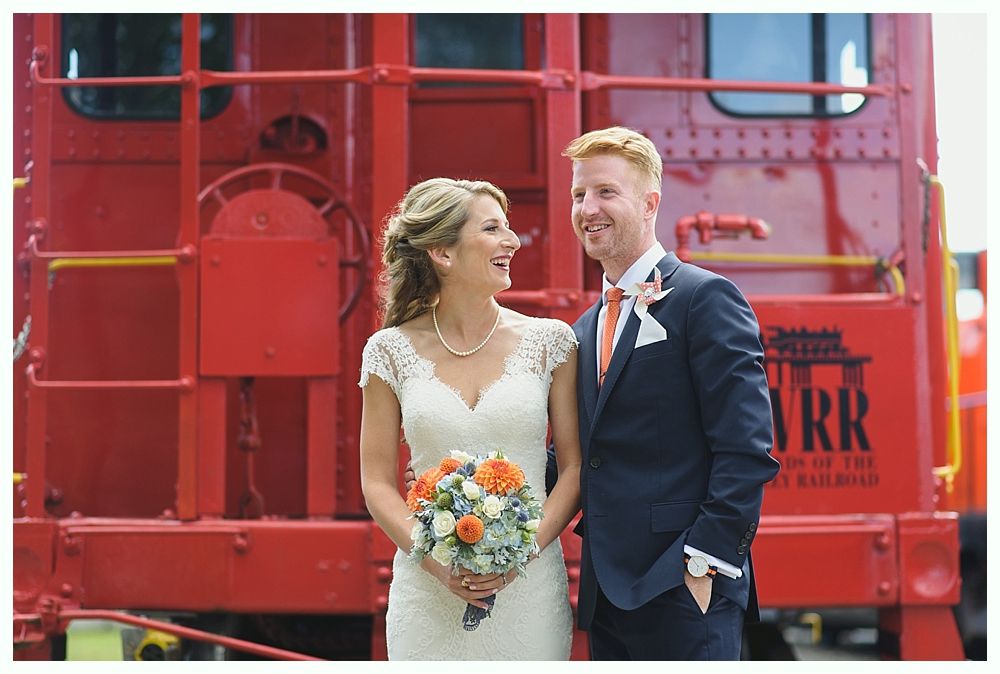 Bride and groom laugh, holding flowers, in front of a red train car.