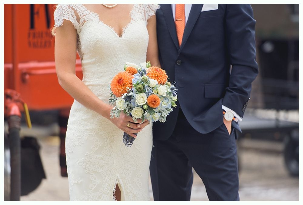 Bride in lace dress holding bouquet, stands next to groom in navy suit, orange tie.