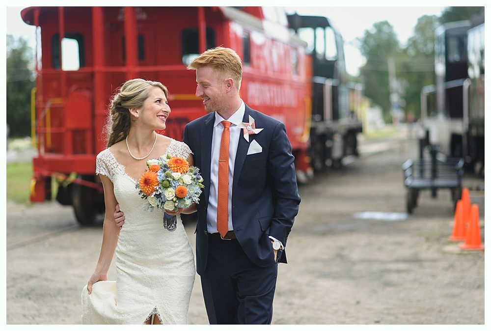 Bride and groom walking, smiling, near a red train car. Bride in white dress, holding flowers, groom in navy suit and orange tie.