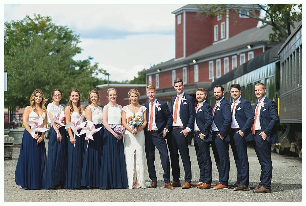 Wedding party posing by train; bride in white, bridesmaids in navy, groomsmen in navy with orange accents, red brick building in background.
