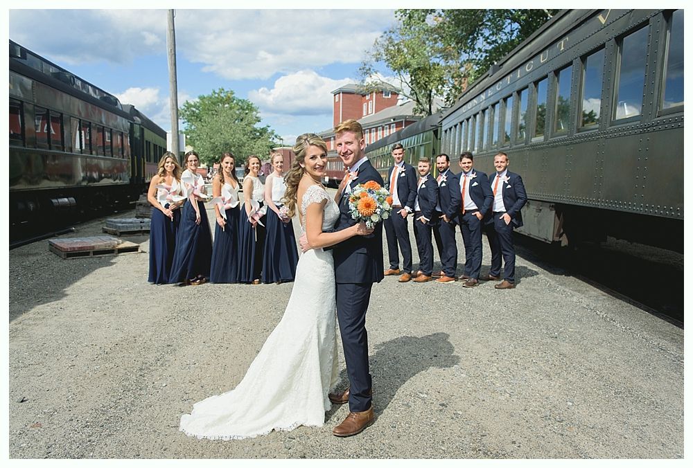 Bride and groom pose with wedding party near vintage train cars. Bride in white gown, groom in suit.