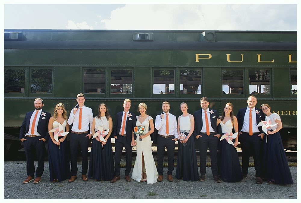 Wedding party poses in front of a green Pullman train car. They wear navy and orange formal attire.