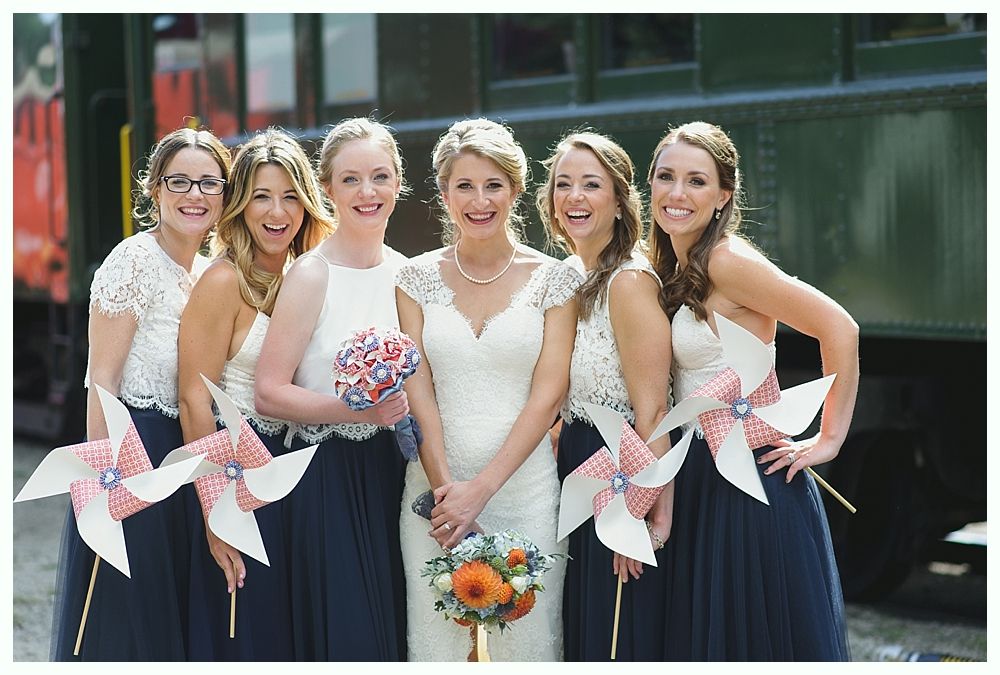 Bride and bridesmaids in navy and white dresses holding flowers and pinwheels in front of a train.