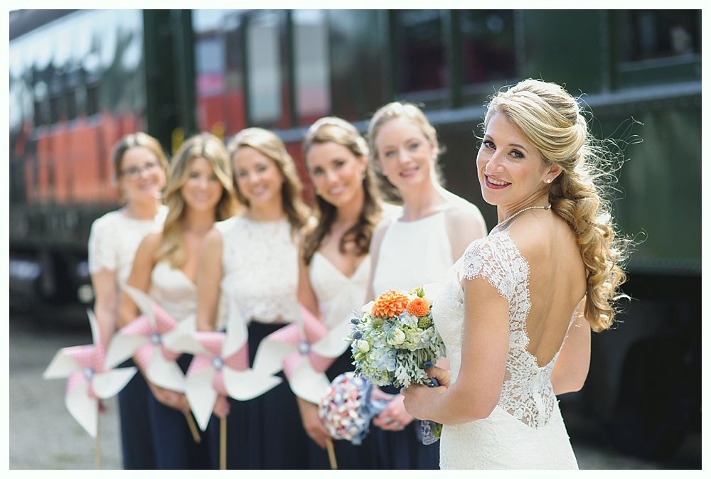 Bride and bridesmaids pose with pinwheels near a train. Bride smiles, wearing a white dress. Bridesmaids hold flowers.