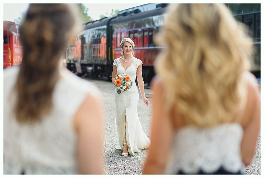 Bride walks toward bridesmaids near train, holding bouquet; sunny day.