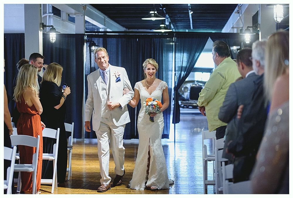 Bride and man walk down aisle. She wears a white dress, carrying flowers. He wears a light suit. Guests watch.