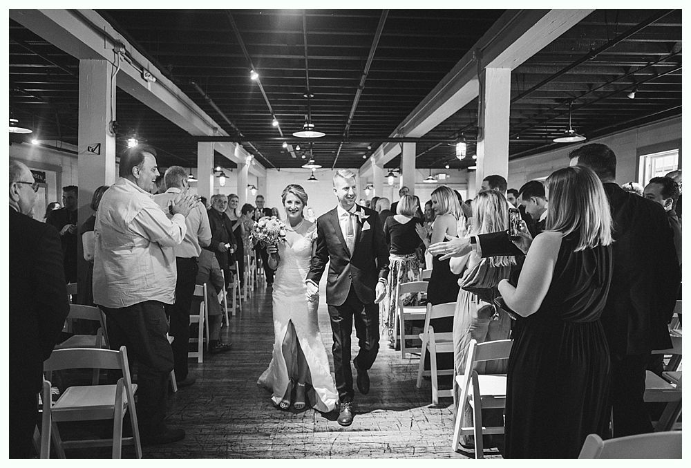 Bride and groom walk down aisle after ceremony, guests watching in indoor venue.