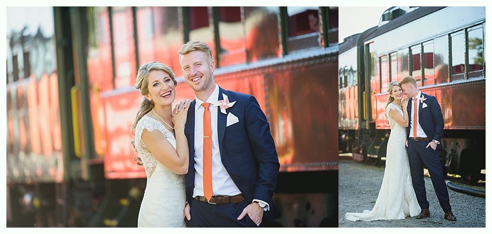 Bride and groom pose by a vintage train. The woman wears a white dress and the man a navy suit with orange tie.