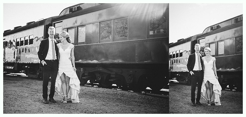 Bride and groom pose in front of a train car. Bride in a wedding dress, groom in a suit. Black and white.