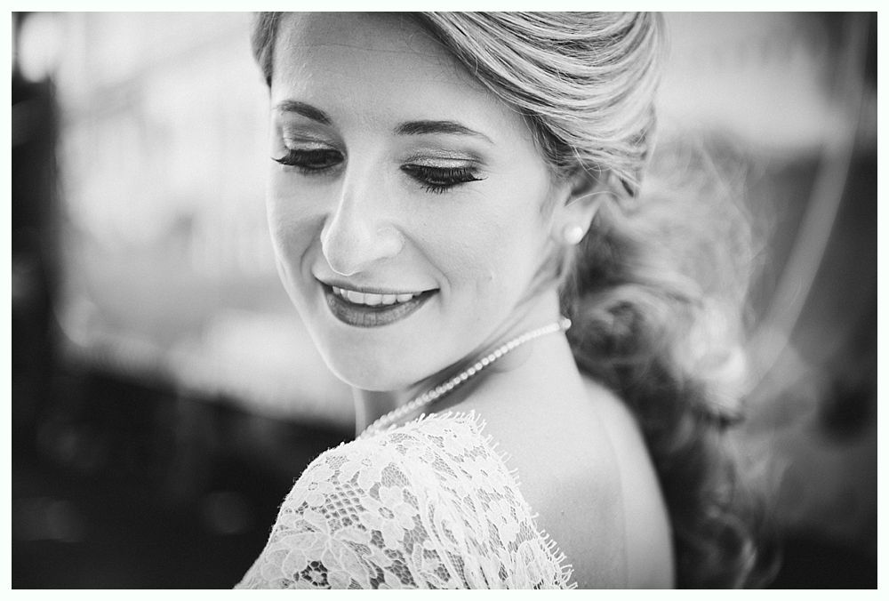 Bride smiles, looking down; lace dress, pearl necklace, updo hairstyle.