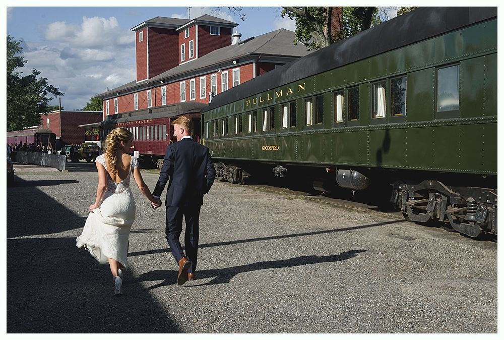 Bride and groom walk hand-in-hand past a green Pullman train car; building in the background.