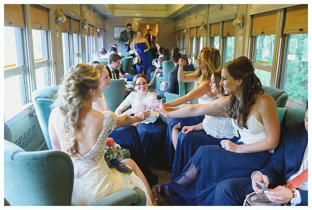 Bridesmaids in navy dresses toast on a vintage train; bride in lace dress seated nearby.