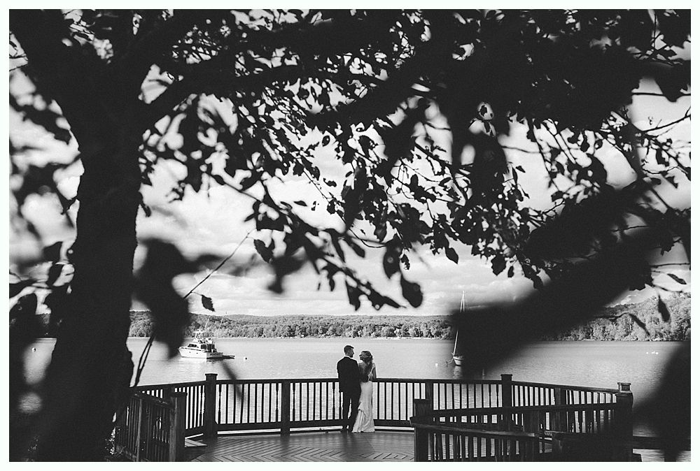 Couple on a wooden deck overlooking a lake, framed by tree branches. Black and white photo.