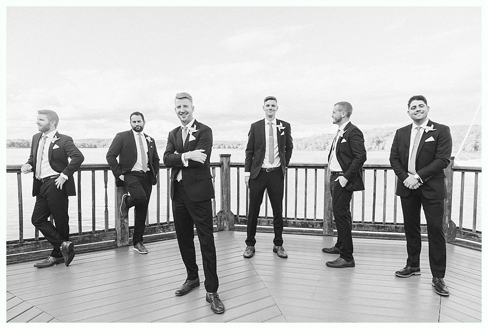 Six men in suits pose on a wooden deck overlooking a body of water.