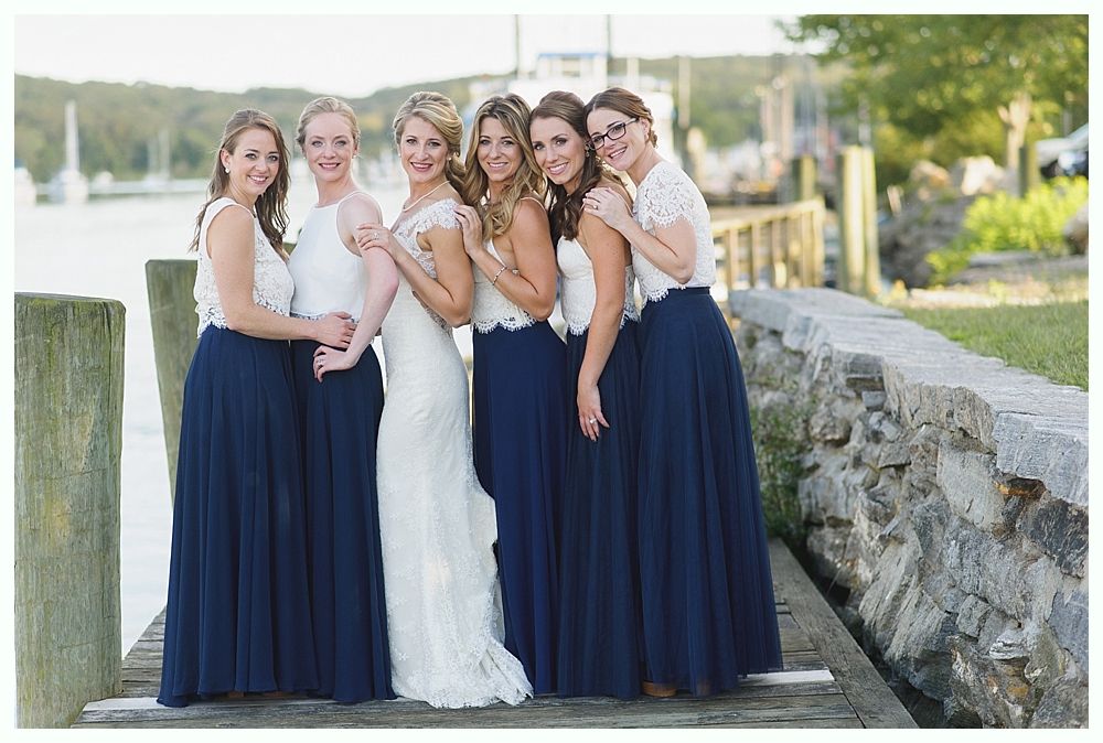 Bridesmaids in navy skirts and white lace tops stand with a bride in a white gown on a dock.