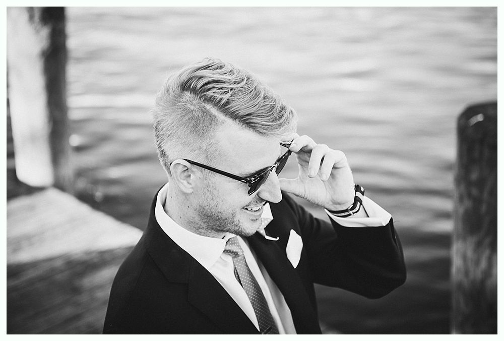 Man in suit and sunglasses adjusts glasses, smiling, near water.