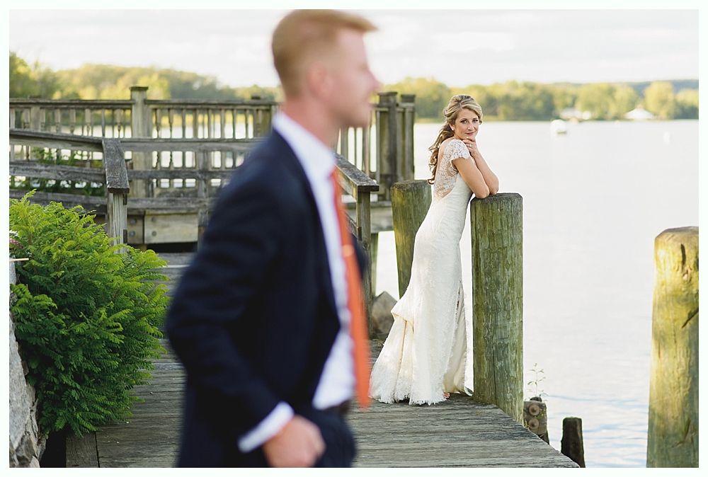Bride in wedding dress leaning on dock, groom blurred in foreground, lake in background.
