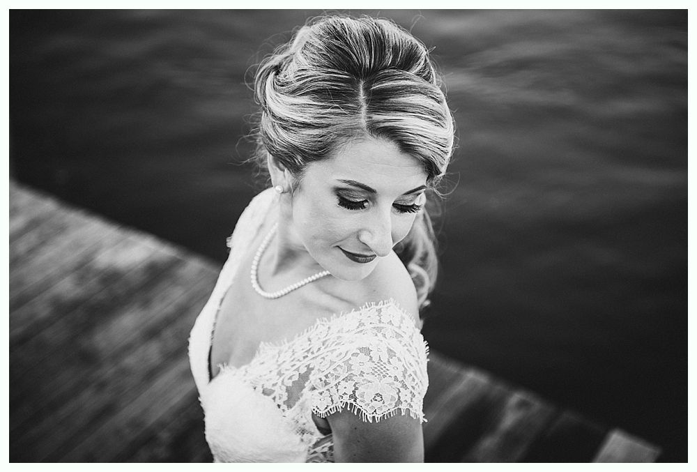 Bride in lace dress, looking down, next to water.