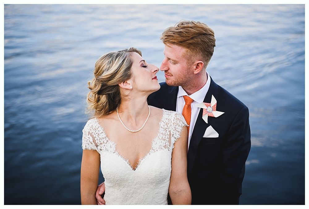 Bride and groom embrace by water. She wears a white lace gown. He wears a suit with an orange tie.