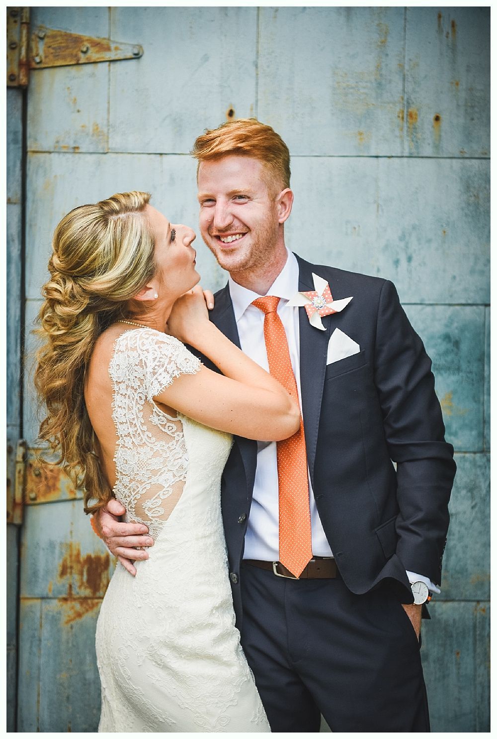 Bride and groom embrace, bride touches groom's face. They smile, wearing wedding attire; blue door background.