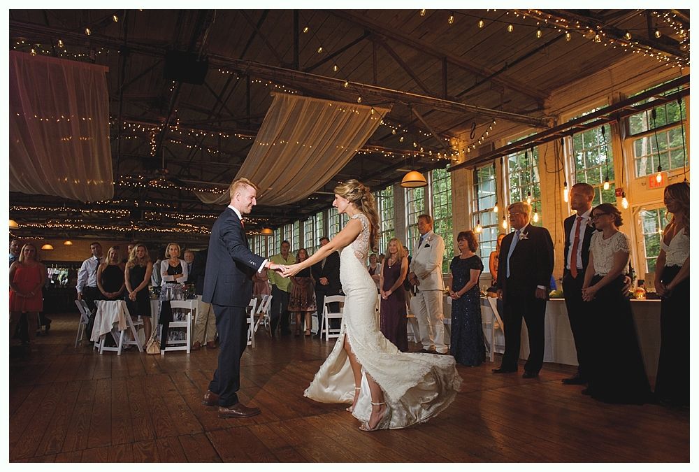 Couple dancing at a wedding reception, holding hands, illuminated by string lights.