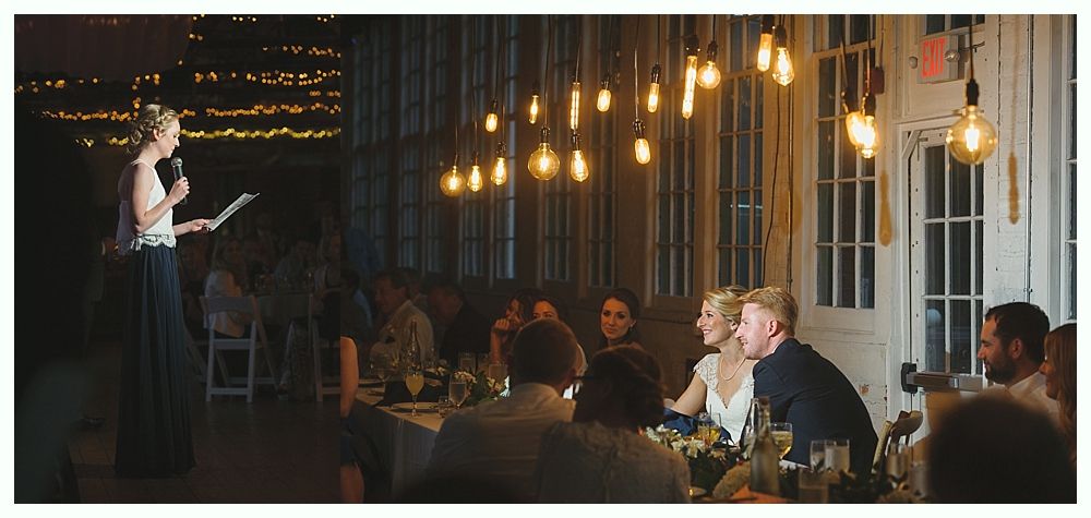 Woman giving a speech at a wedding, illuminated by hanging lights, with the bride and groom seated at a table.