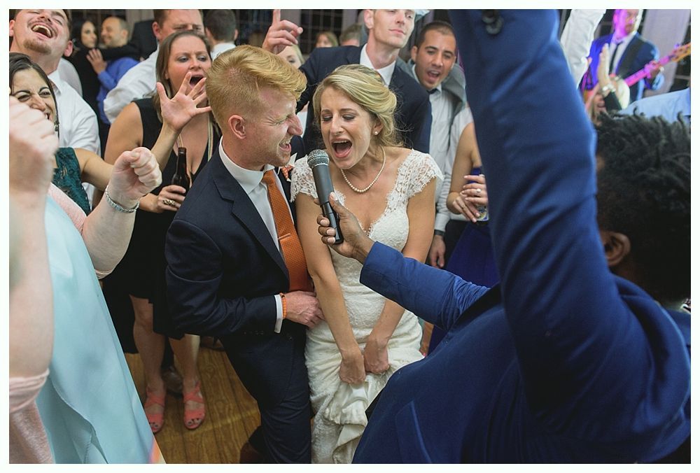Bride and groom singing into microphone, surrounded by cheering guests at a wedding reception.