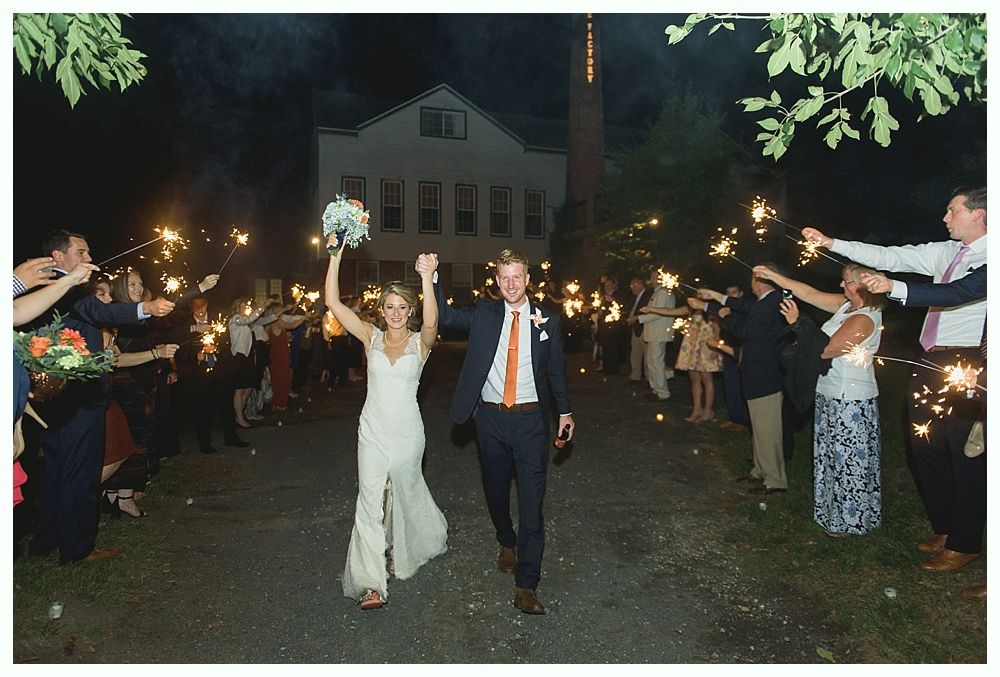 Bride and groom exit reception with sparklers held by guests at night. Building in background.