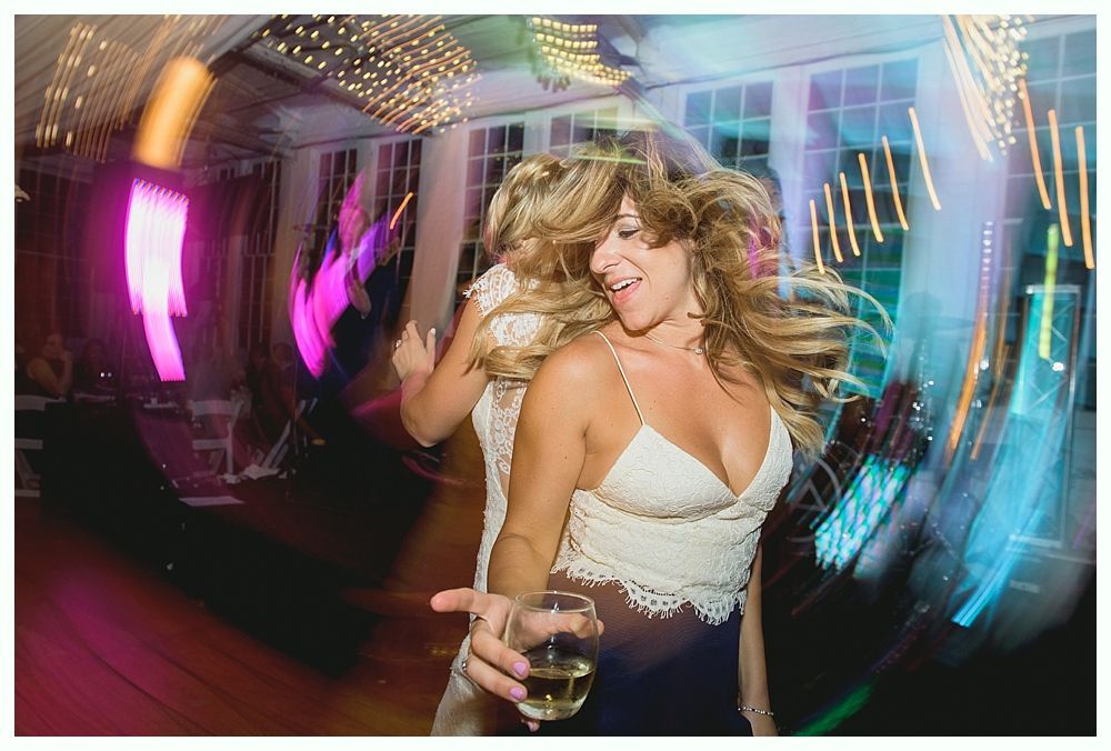 Two women dancing at a party, one holding a drink. Bright lights blur around them.