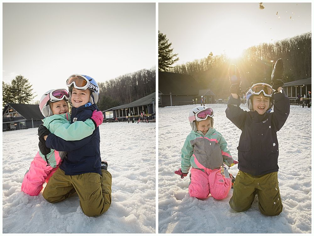 Two children in ski gear play in the snow. One hugs the other, the second throws snow in the air.