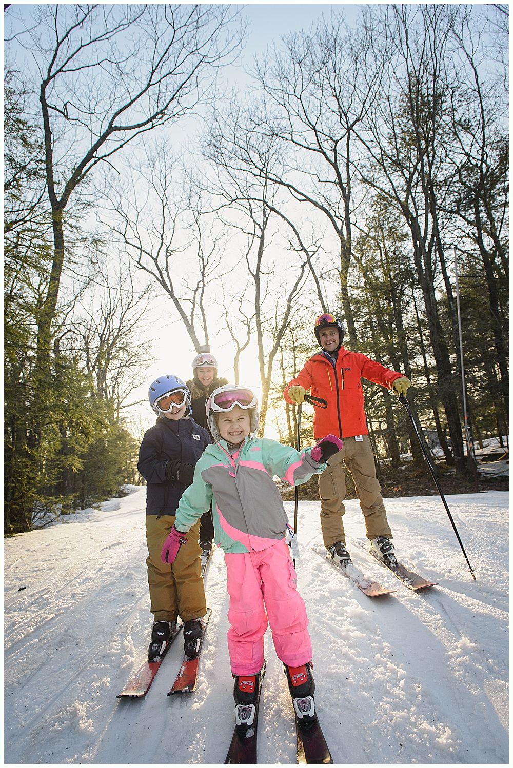 Family on skis, smiling at camera, snowy slope, trees and sunlight in background.
