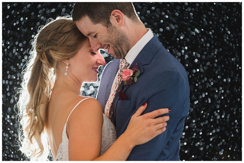 Bride and groom embrace, touching foreheads. The bride wears a white dress, the groom a blue suit. Sparkling dark background.