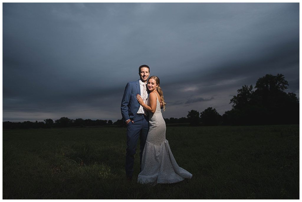 Couple in wedding attire embrace in a field, dark storm clouds overhead.