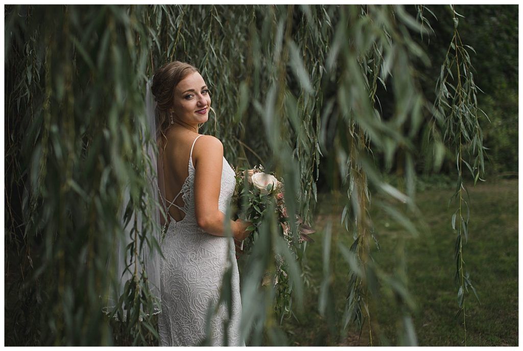 Bride in a white beaded wedding dress, holding flowers, under willow tree branches; smiling.