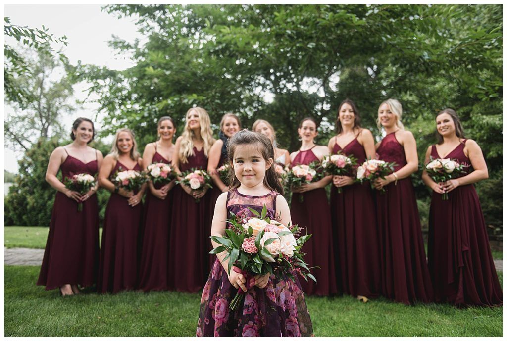 Flower girl stands before bridesmaids in burgundy dresses, holding bouquets outdoors.