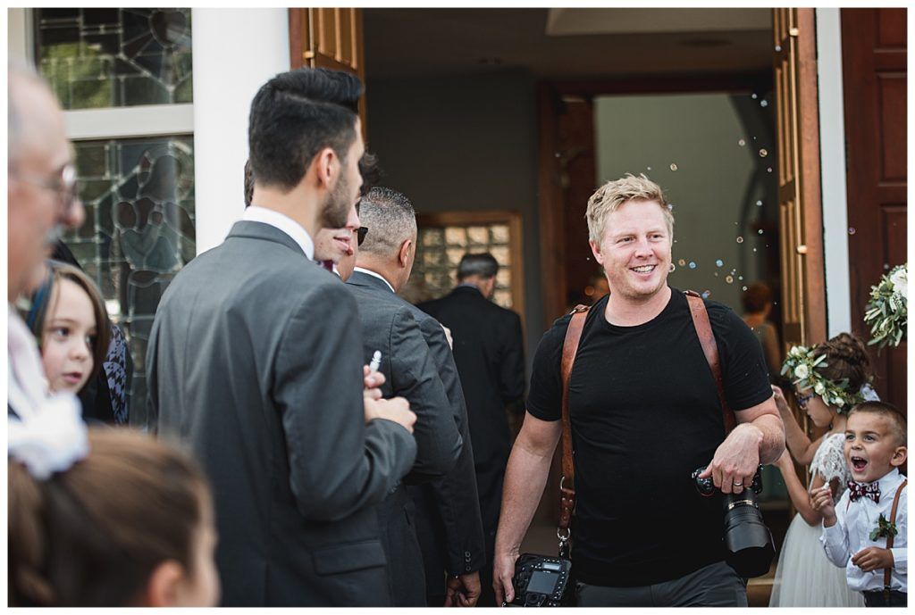 Wedding guests exiting a building, photographer smiling, confetti in the air.
