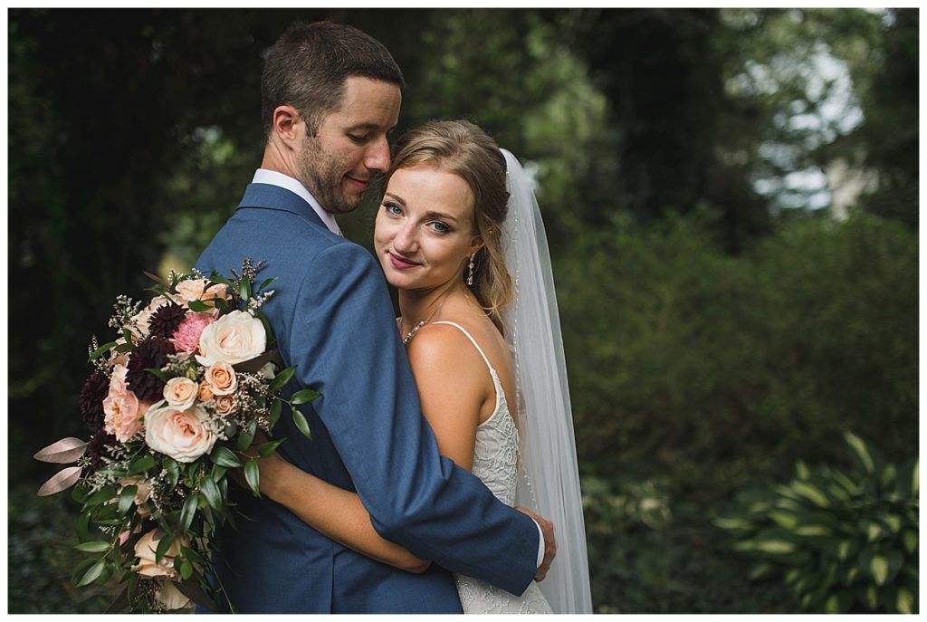 Bride and groom embrace outdoors; she holds a bouquet, he wears a blue suit; both smiling.