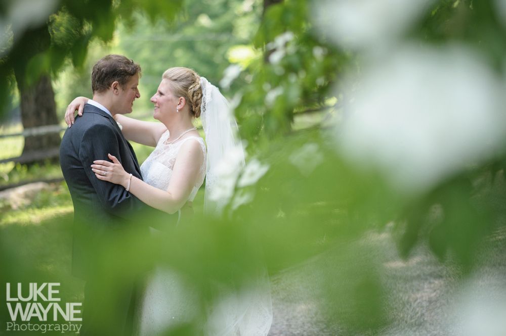 Couple embracing outdoors, bride in white dress, groom in suit, surrounded by greenery.