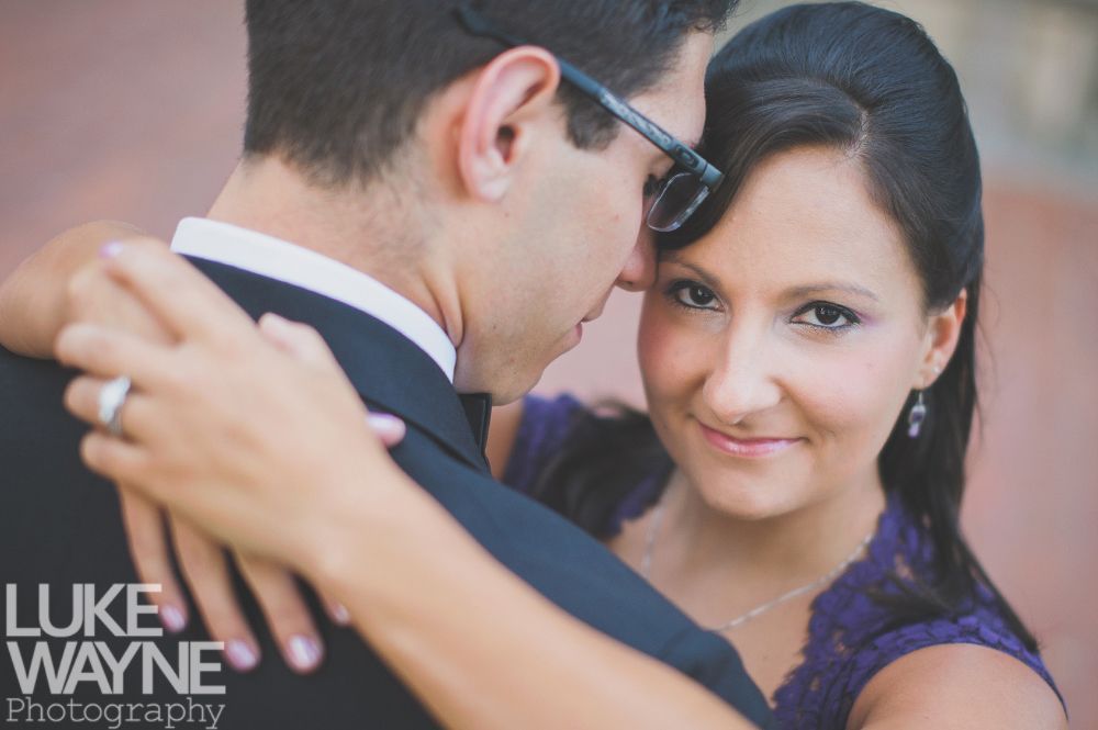 Couple embracing, woman looks at camera, wearing purple dress, man wearing suit, glasses.