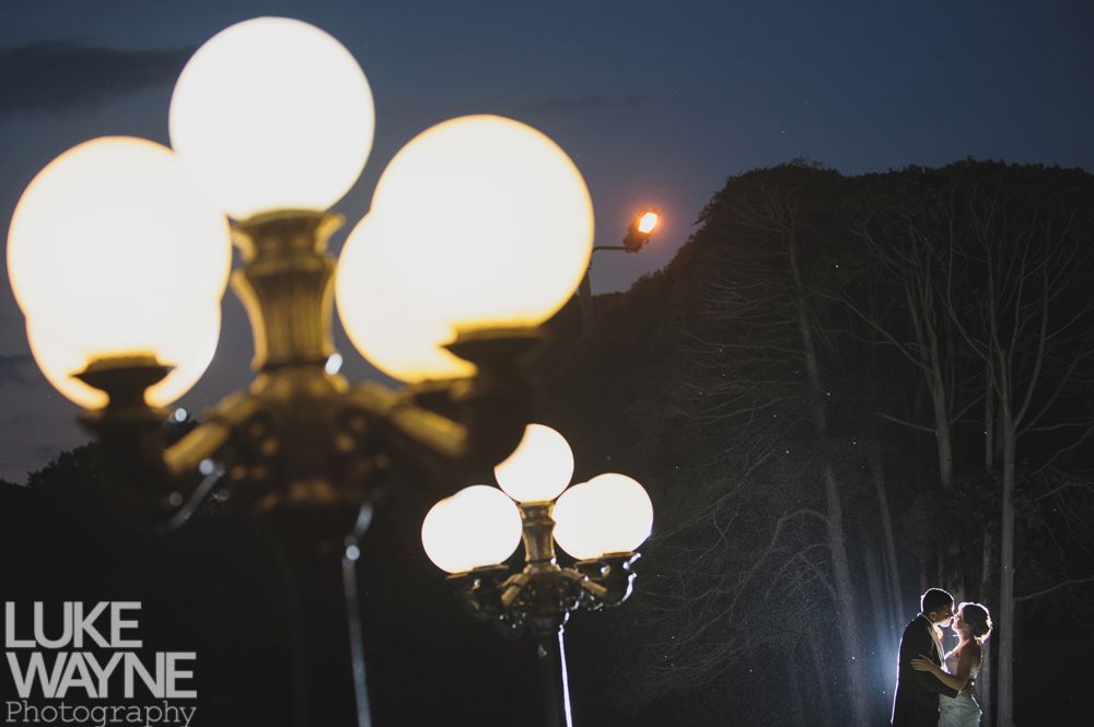 Couple kissing near trees at night with glowing streetlights in foreground.