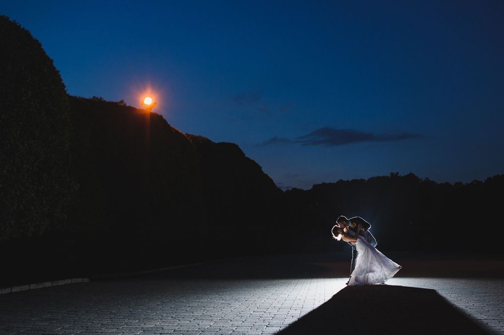 Bride and groom embrace, lit by spotlight against dark night sky and hillside.