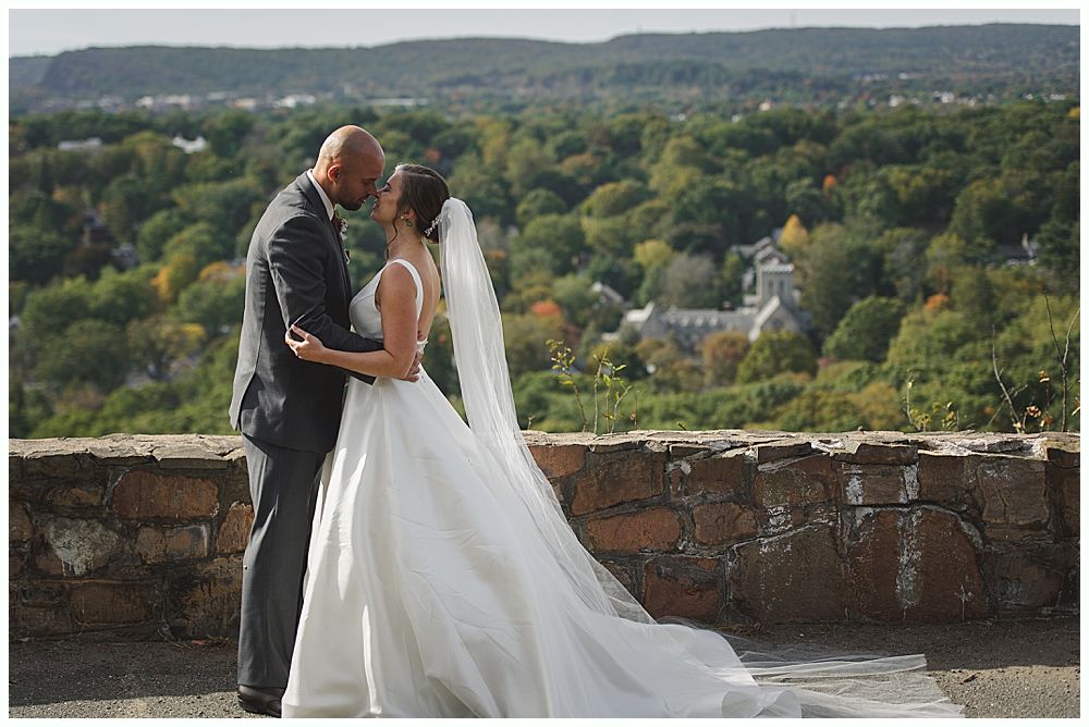People seated at outdoor ceremony. Elderly woman in beige coat, couple in formal attire, others in background.
